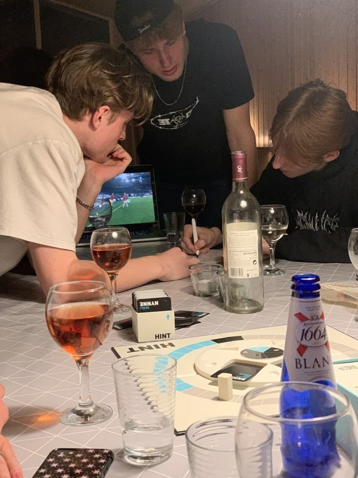 This may contain: three men sitting at a table with wine glasses and bottles on it, looking at something in front of them