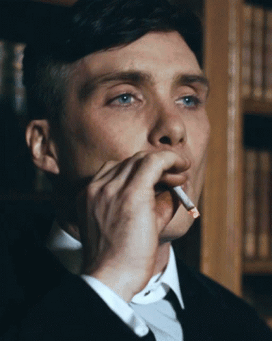 a man in a suit is smoking a cigarette in front of a bookshelf