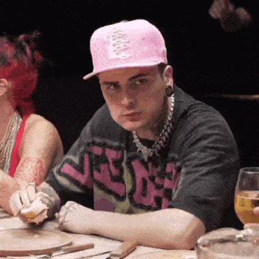 a man wearing a pink ny hat sits at a table eating a sandwich