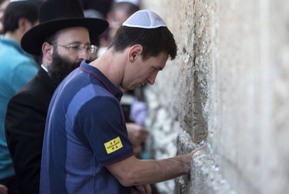 This may contain: a man wearing a blue shirt and black hat standing next to a stone wall with writing on it