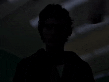 a man in a red shirt is standing in a dark room with a ceiling fan behind him